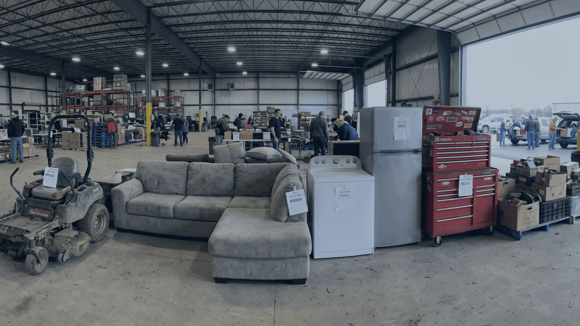 Warehouse interior with a lawnmower, sofa, appliances, and people browsing various goods.