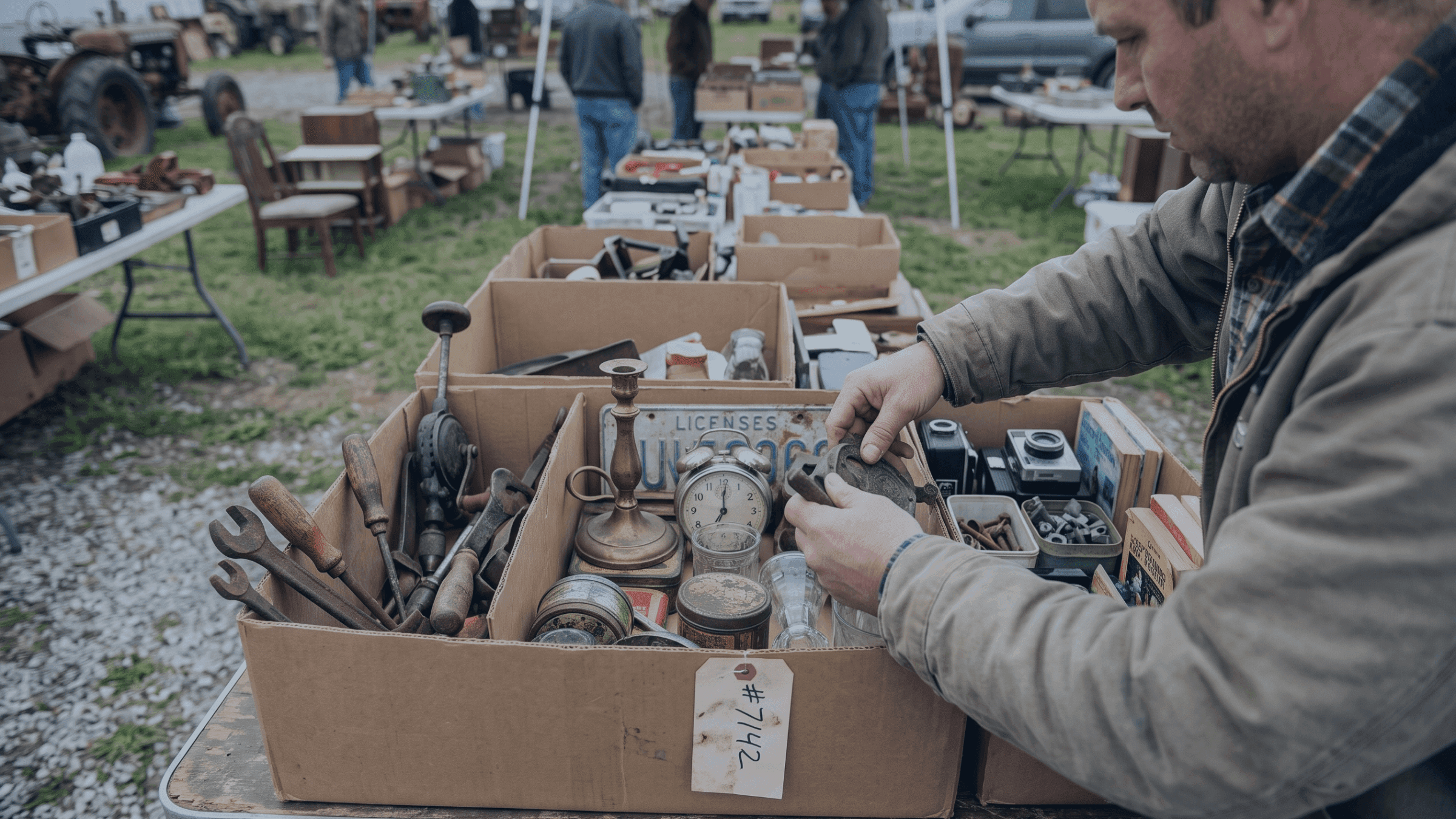 Man inspects old tools and antiques in a cardboard box at an outdoor flea market.